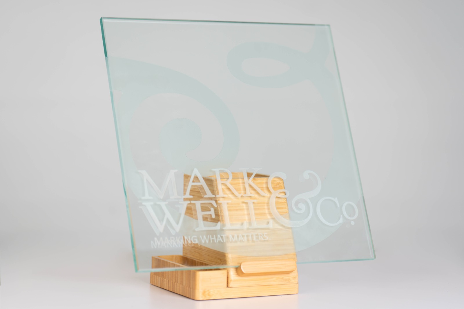 Sand-carved glass panel bearing the MarkWell and Co. mark and the line Marking What Matters, standing on a bamboo base.